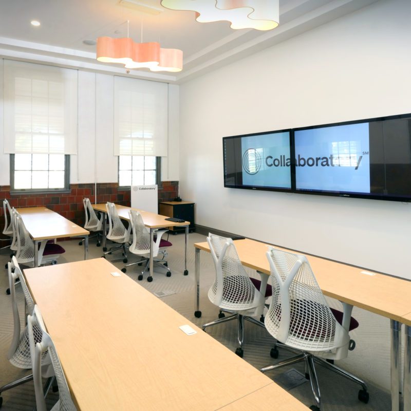 Picture of the Classroom. Shows rolling chairs, tables, windows, and two 70-inch MondoPads (smart-board touchscreens)TV screens.