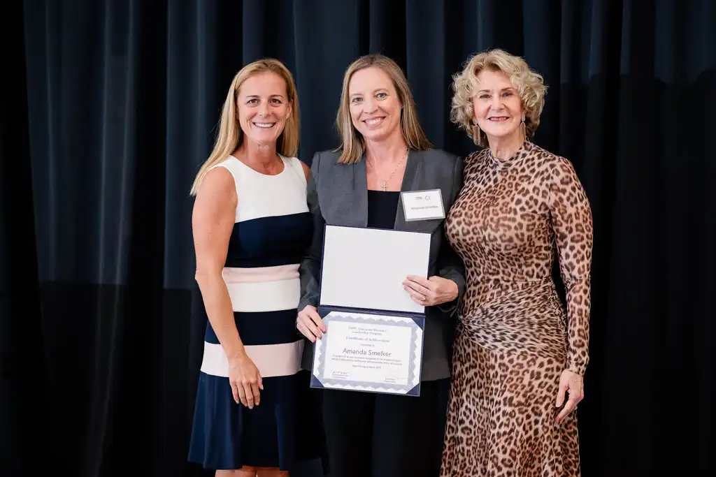 Three women stand on a stage with a dark curtain backdrop, the center woman holding a certificate and smiling.