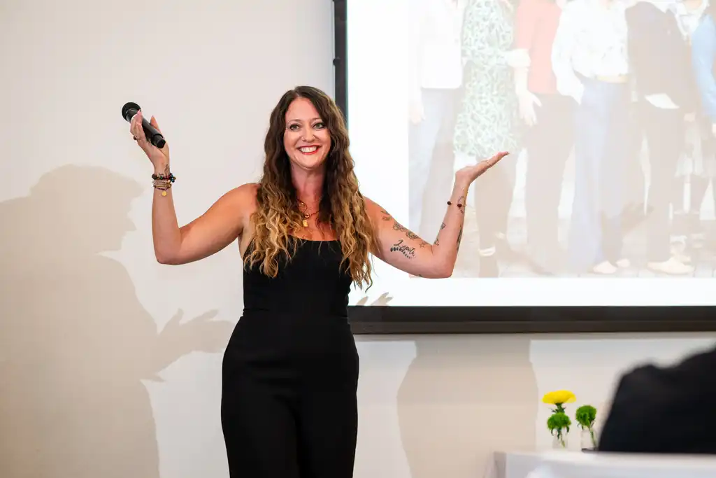 Smiling woman in a black outfit stands on a stage, holding a microphone, with a projector screen and small flowers in the background.