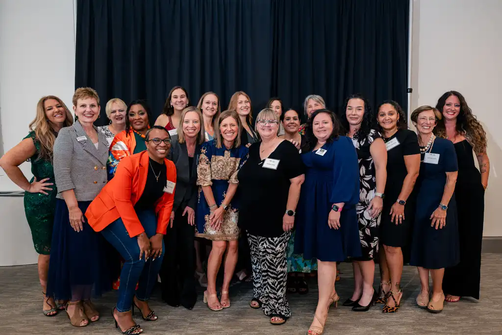 A diverse group of women in dresses and business attire posing together for a group photo indoors.