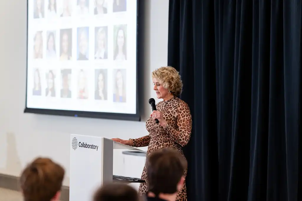 A woman in a leopard-print dress speaks at a podium during a presentation, with a screen displaying a grid of headshots behind her.