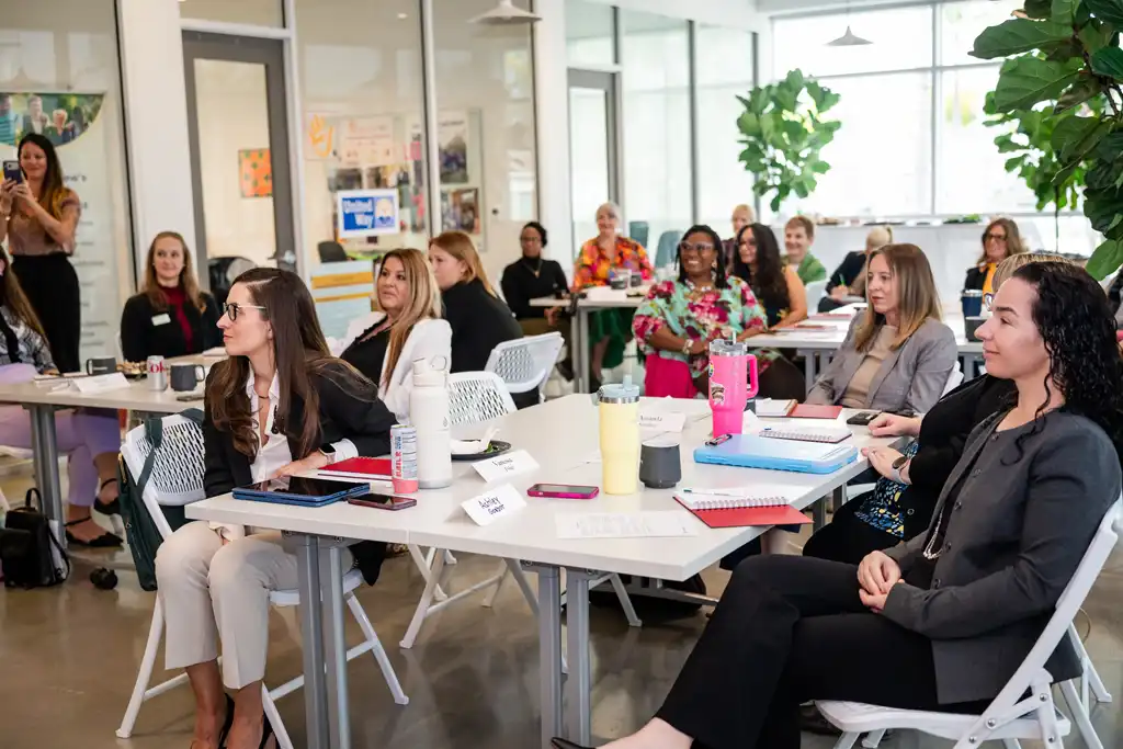 Group of women seated at white tables in a bright open room, attentively listening to a workshop presentation.