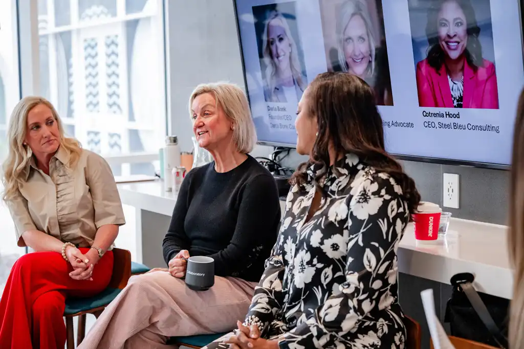 Three women sit on chairs in a bright studio during a panel discussion, with portrait images on screens in the background.