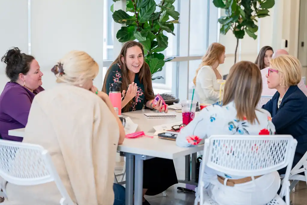 A group of people seated around a table in a bright room with large leafy plants, chatting and smiling.