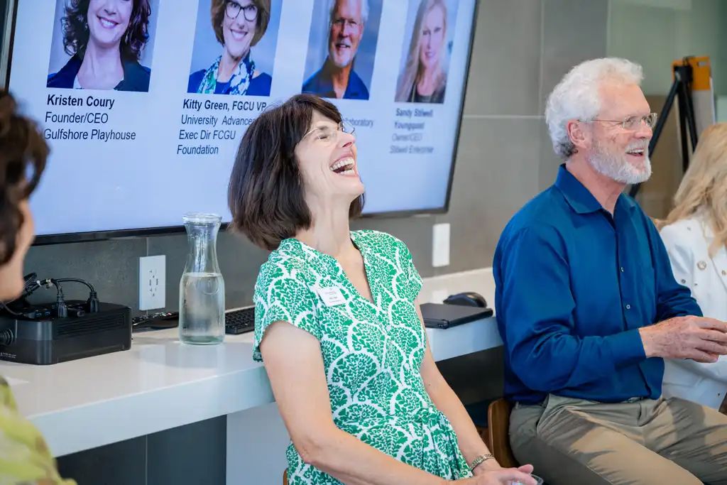 A woman in a green patterned dress laughs while seated at a panel beside a man in a blue shirt, with a screen showing headshot portraits in the background.