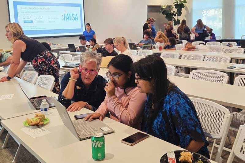 Three people collaborate on a laptop at a long white table in a bright conference room, with snacks and drinks nearby as others work in the background.