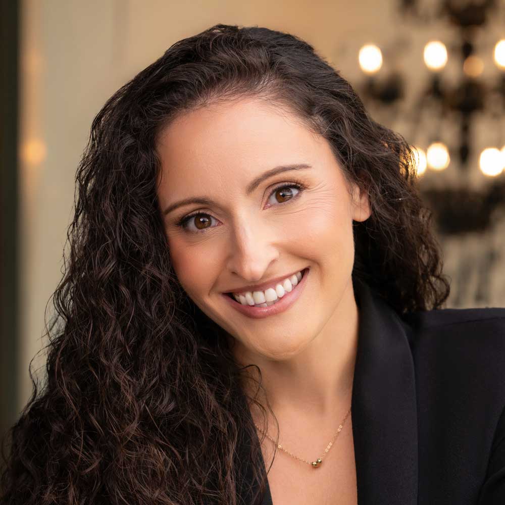 Andrea Cappuccilli Smiling woman with long curly hair wearing a black blazer and a delicate necklace, indoors with warm lights in the background.